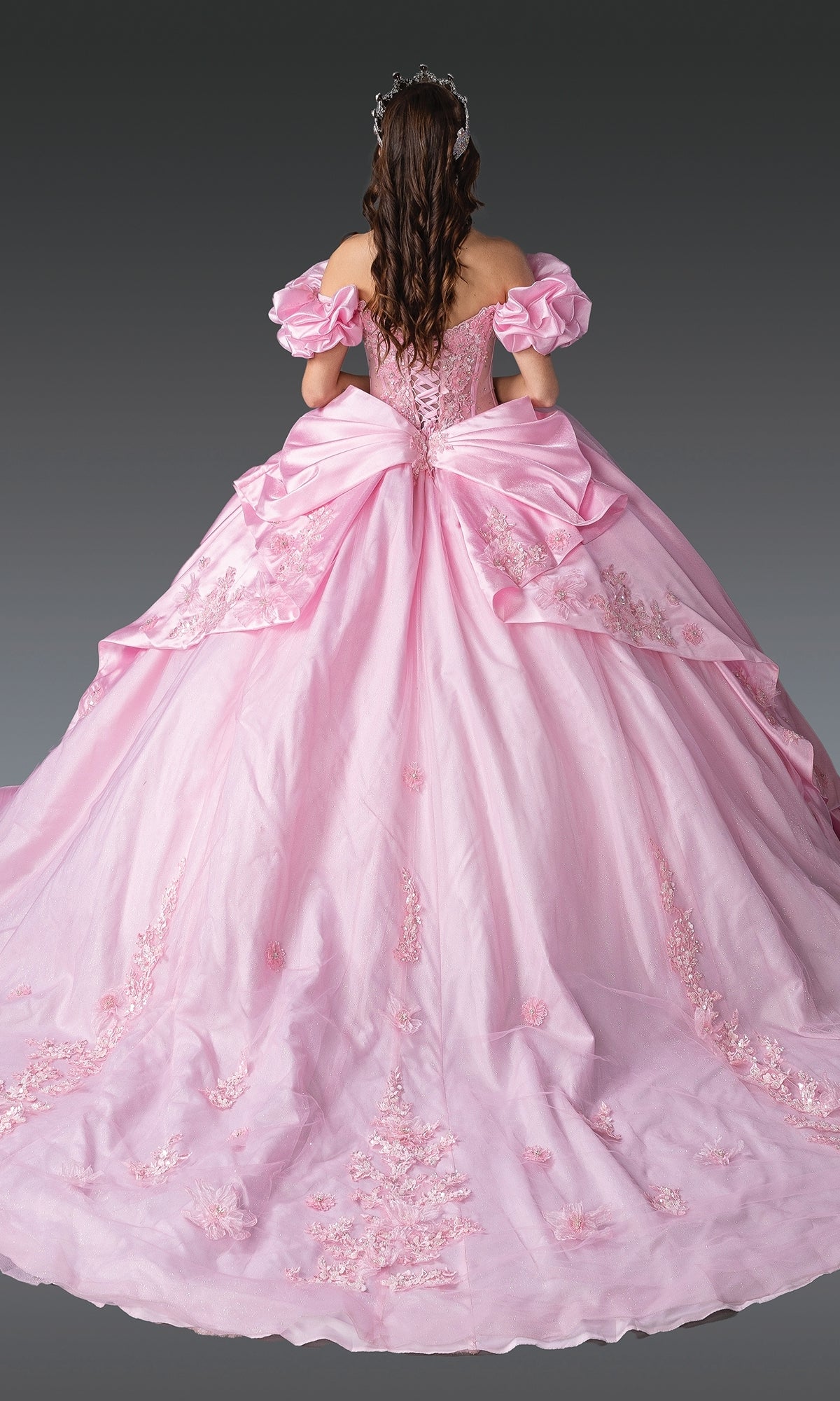 A woman with long brown hair faces away, modeling the Quinceanera Dress 1994 by Dancing Queenโa light pink gown with a large bow, floral embroidery, and puffed sleevesโset against a plain gray background.